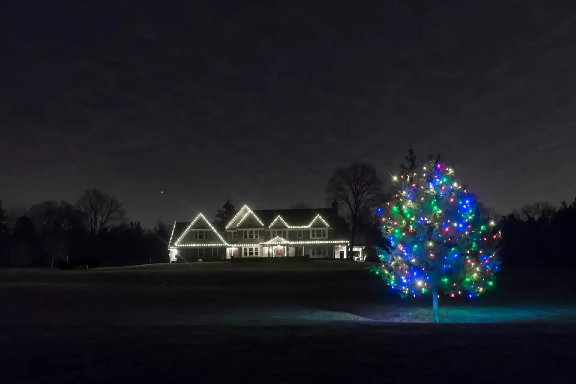 House and large tree outlined with white and multicolored holiday lights at night