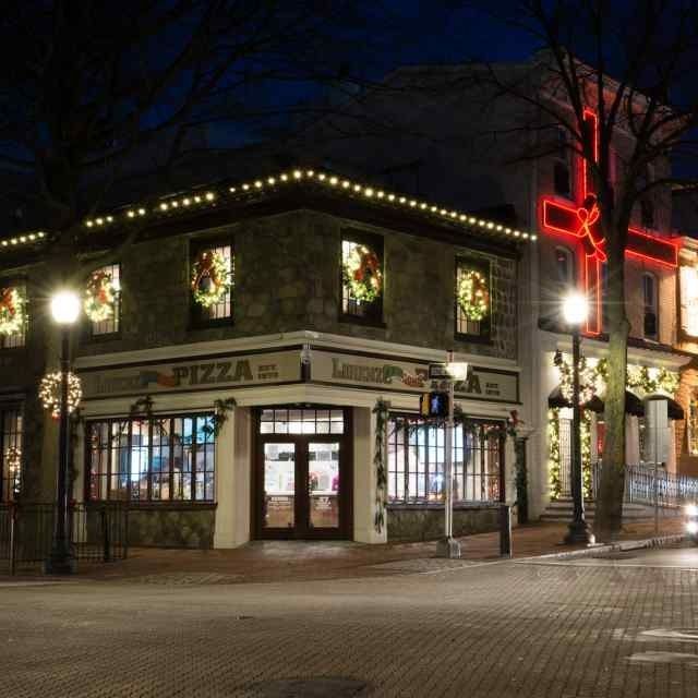 Nighttime corner pizza restaurant with lit windows, holiday lights, and a red neon sign