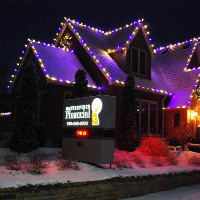 Night-lit house with purple and yellow holiday lights and a glowing sign in snowy front yard