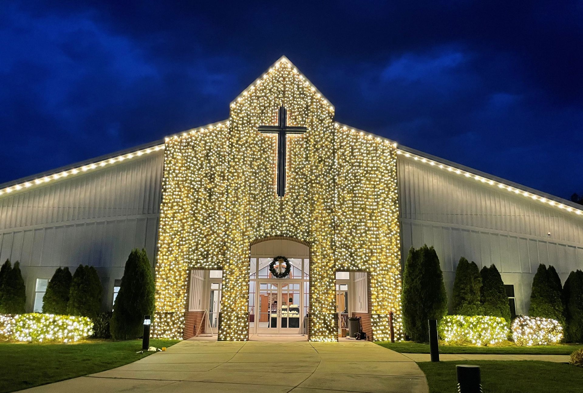 Church entrance at night, covered in warm lights with a large cross above the doors.