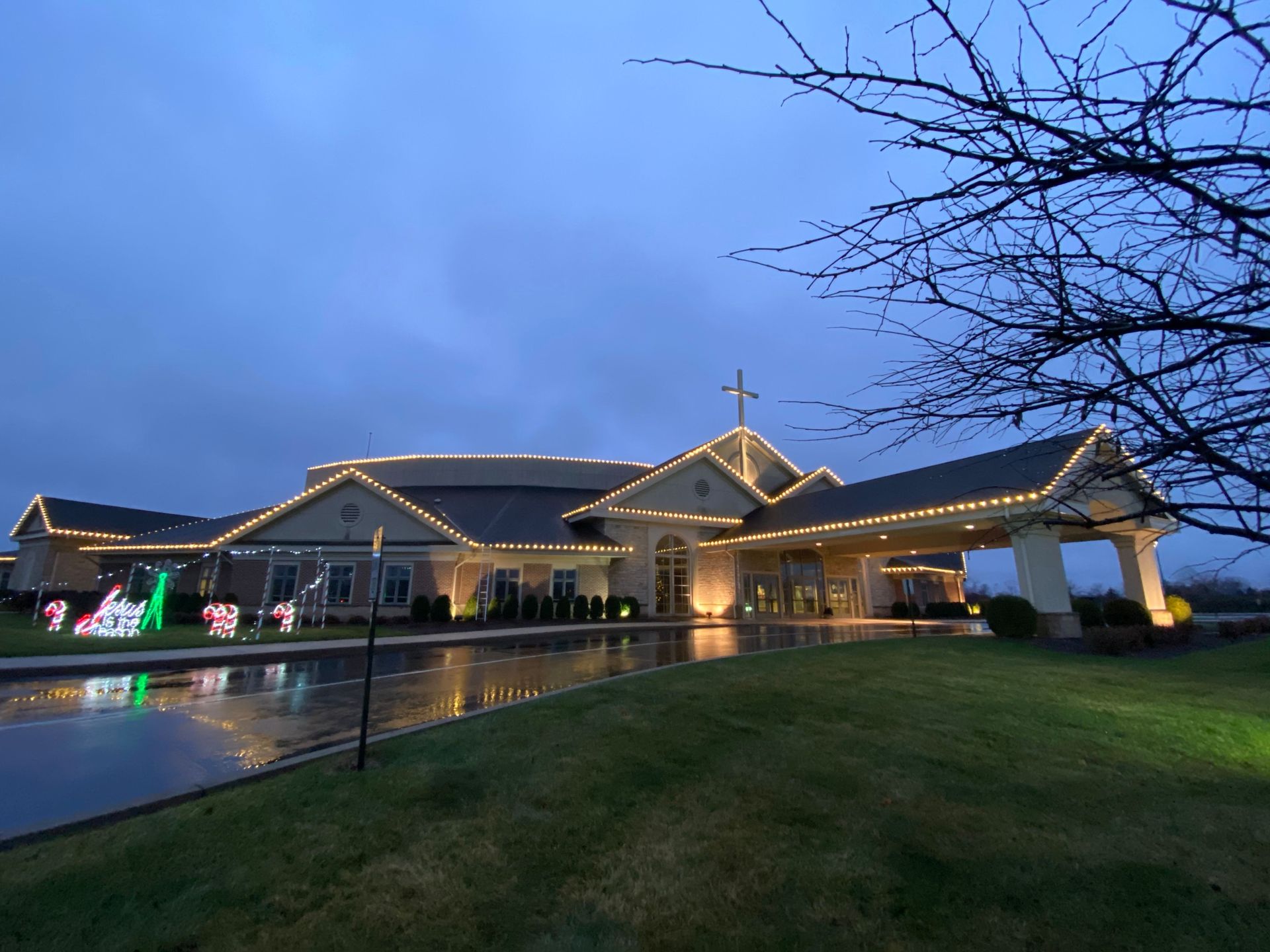 Church building at dusk with warm lights, a cross on the roof, and a wet lawn under a blue sky