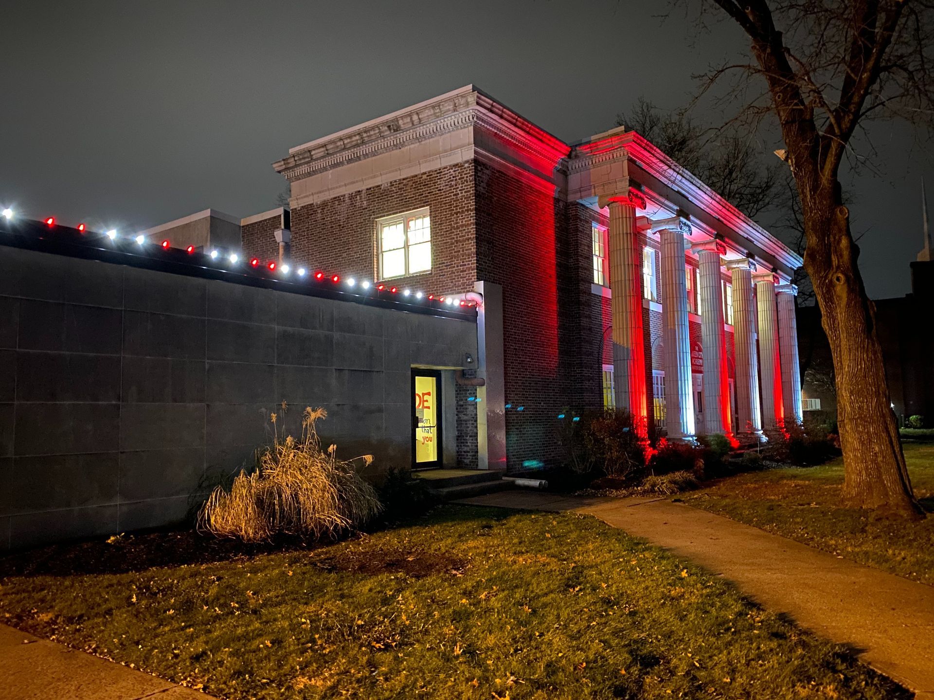 Brick building lit red at night beside a sidewalk and grass, with a tree in the foreground.