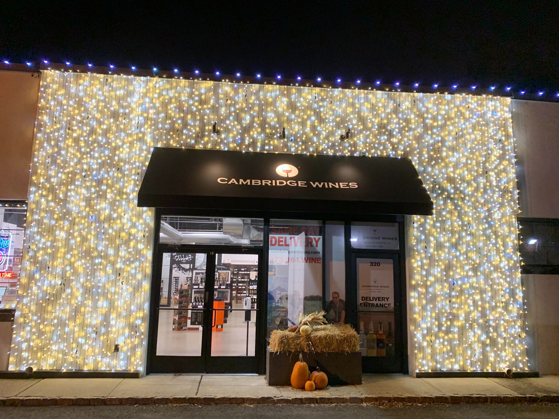 Storefront wrapped in white and gold lights at night, with a black awning and pumpkins by the entrance