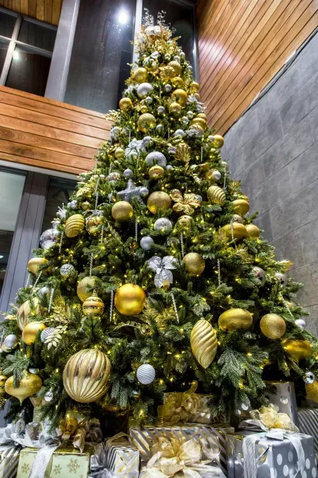 Tall decorated Christmas tree with gold and silver ornaments and wrapped gifts in a modern lobby