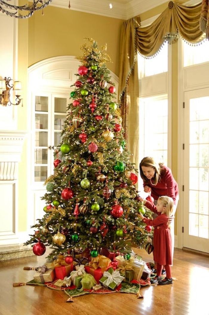 Woman in red decorating a Christmas tree in a bright room with large windows and gold drapes