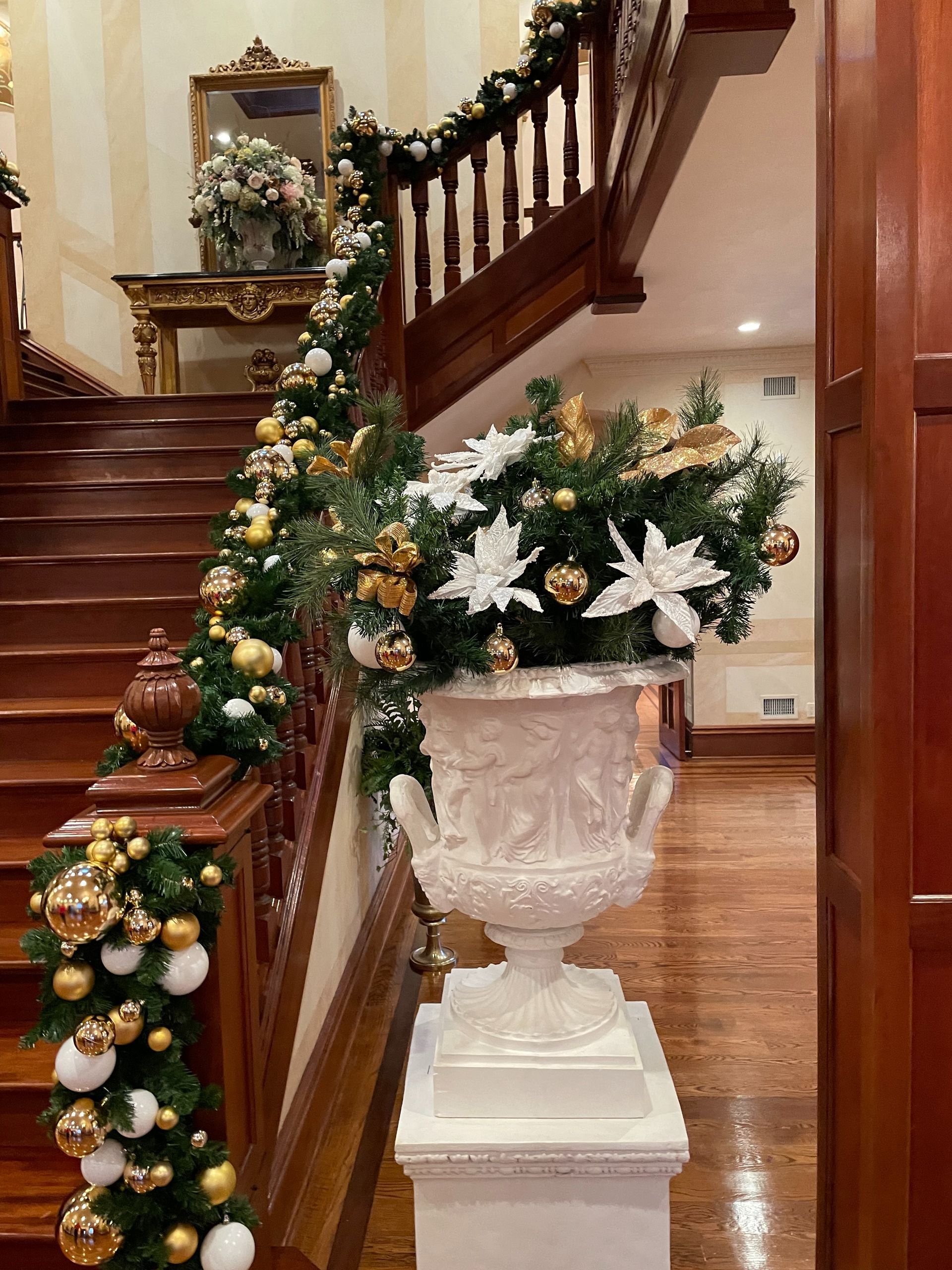 Holiday-decorated staircase with garland, white poinsettias, and a large urn planter in a polished foyer