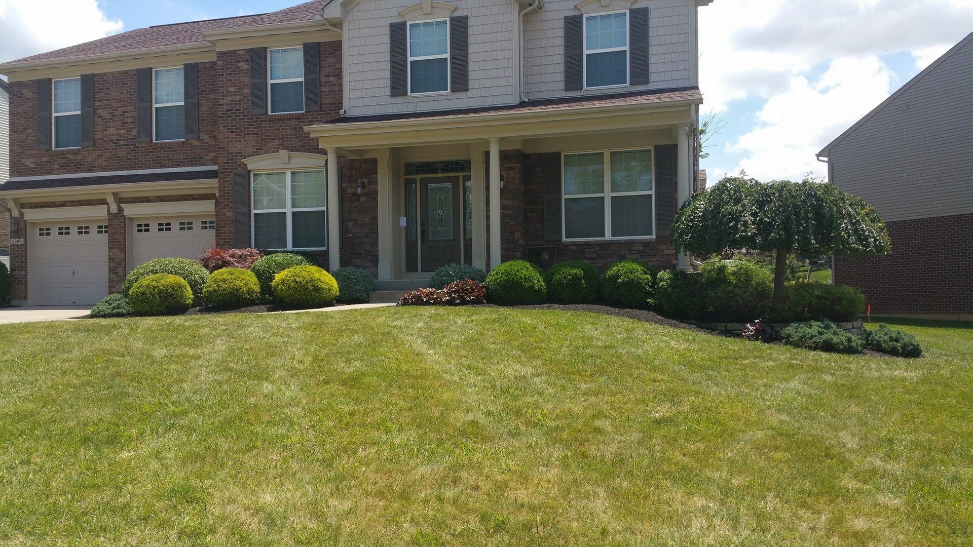Two-story suburban brick house with a front porch, garage, and sloped green lawn