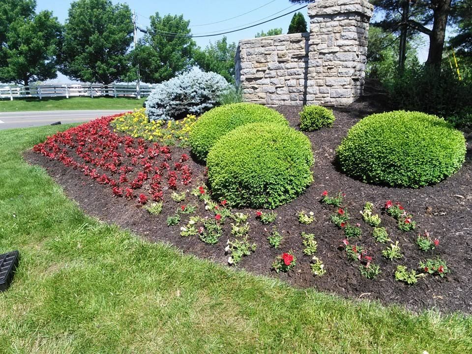 Colorful flower bed with red blooms, green shrubs, and a stone pillar beside a grassy lawn.
