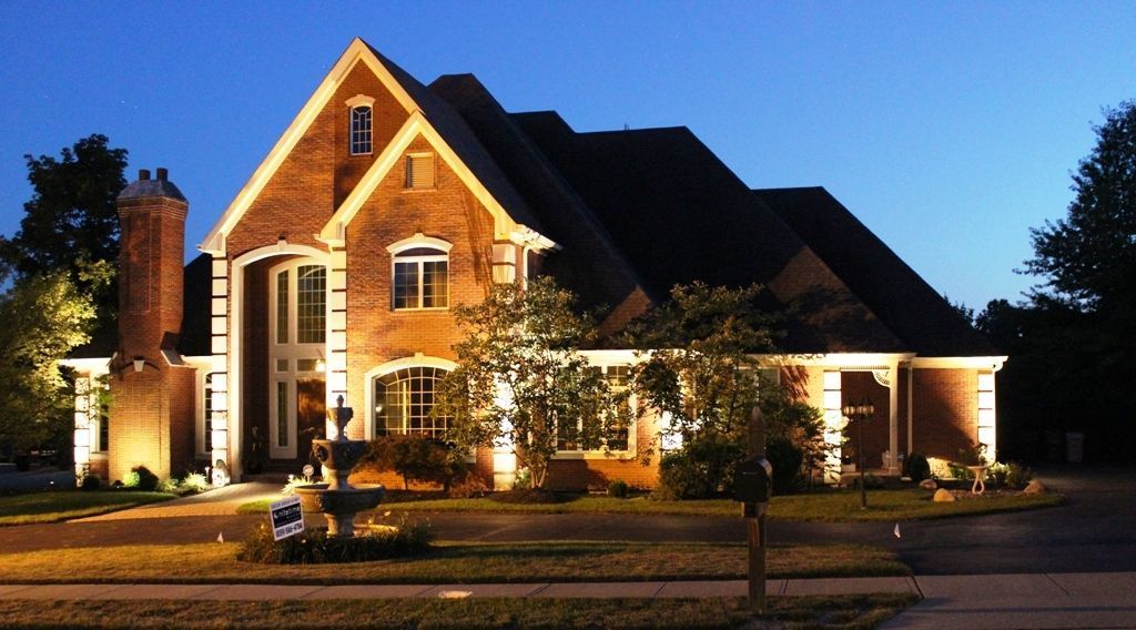 Illuminated brick house with steep roof at night, lit by warm outdoor lights and surrounded by trees.