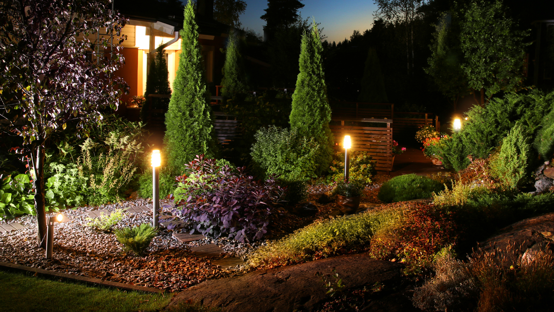 Lit landscaped garden at dusk with trimmed shrubs, trees, and glowing pathway lights