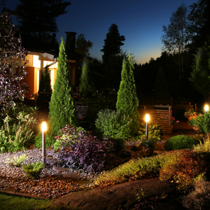 Lit landscaped garden at dusk with evergreens, flower beds, and glowing path lights.