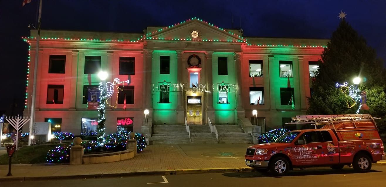 Historic building lit red and green for the holidays, with a decorated vehicle parked in front.