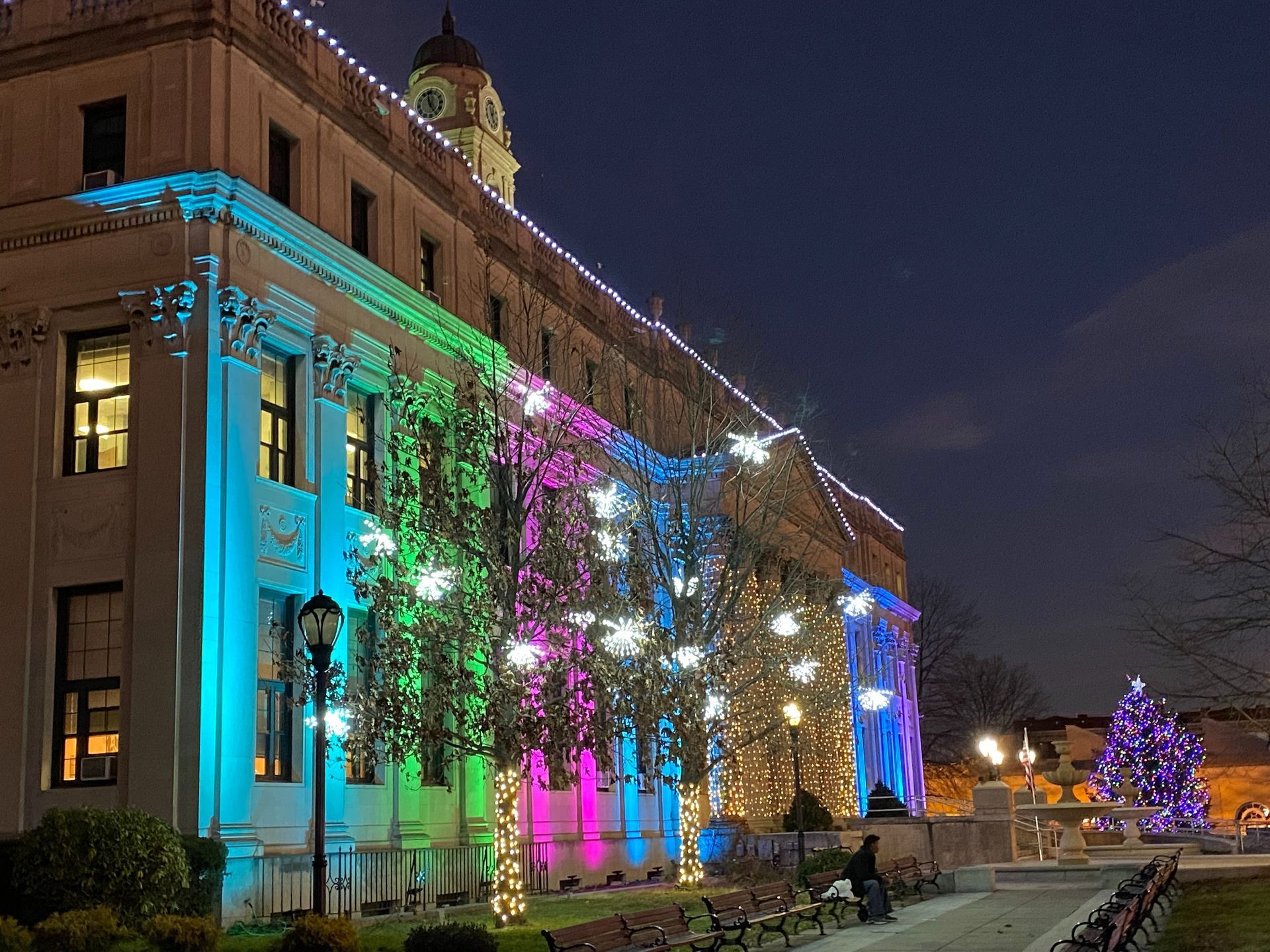 Historic building lit with rainbow lights at night, with a decorated Christmas tree nearby