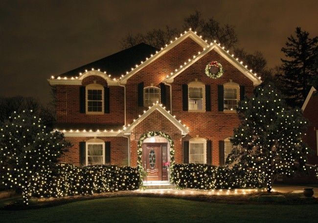 Brick house decorated with warm white Christmas lights, wreaths, and lit bushes at night