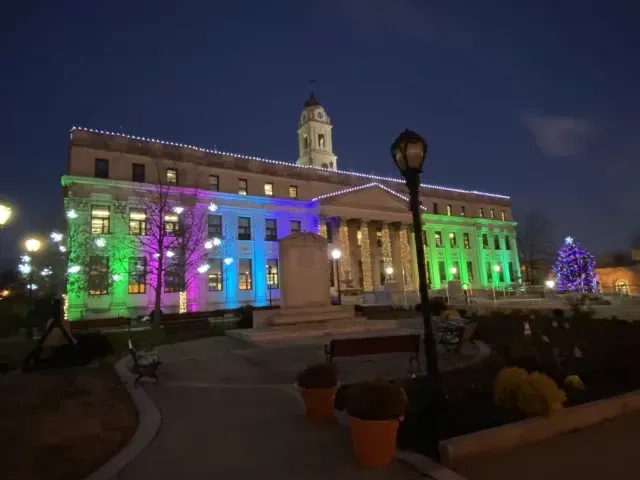 Lit building lit in rainbow colors at night, with a clock tower and plaza in front