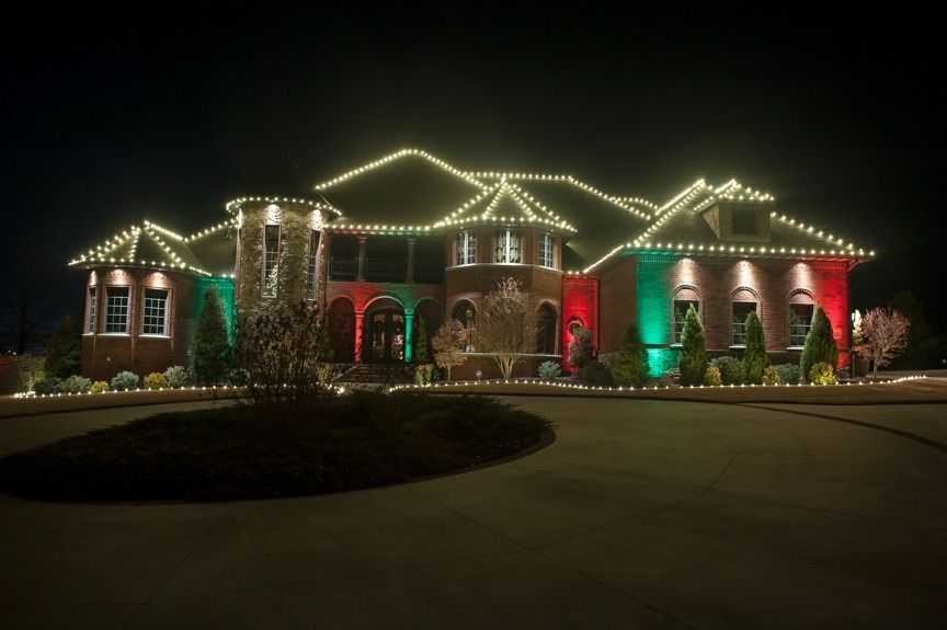 A large house lit with white and red Christmas lights at night, with a dark driveway in front.