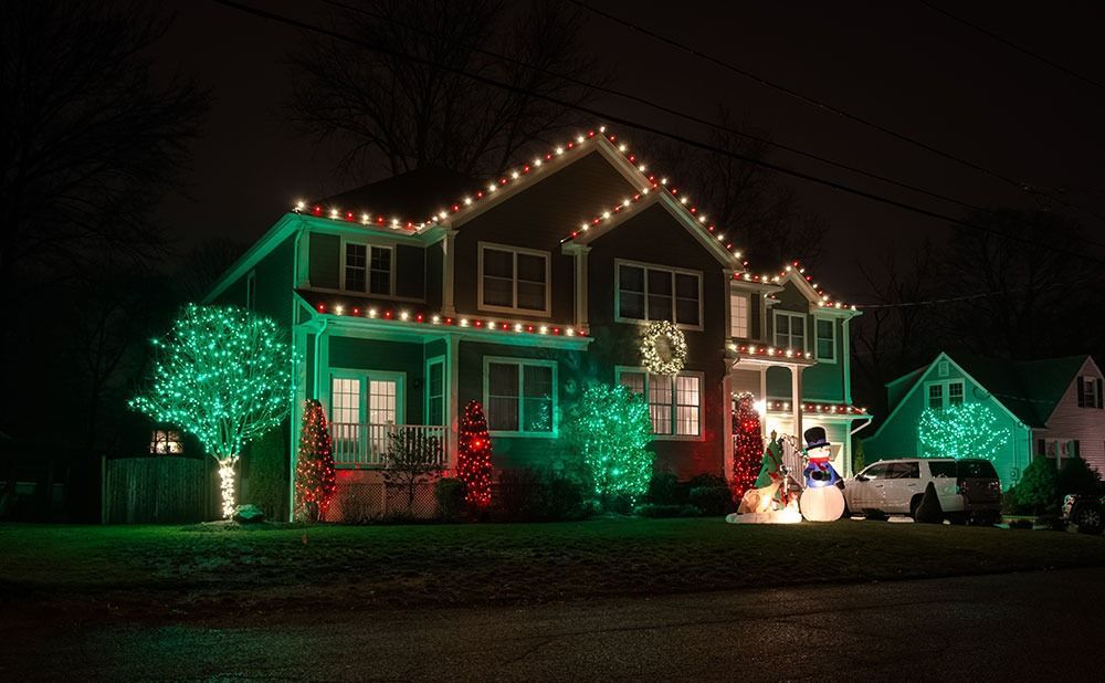 House decorated with green and white Christmas lights at night, with lit trees in the yard