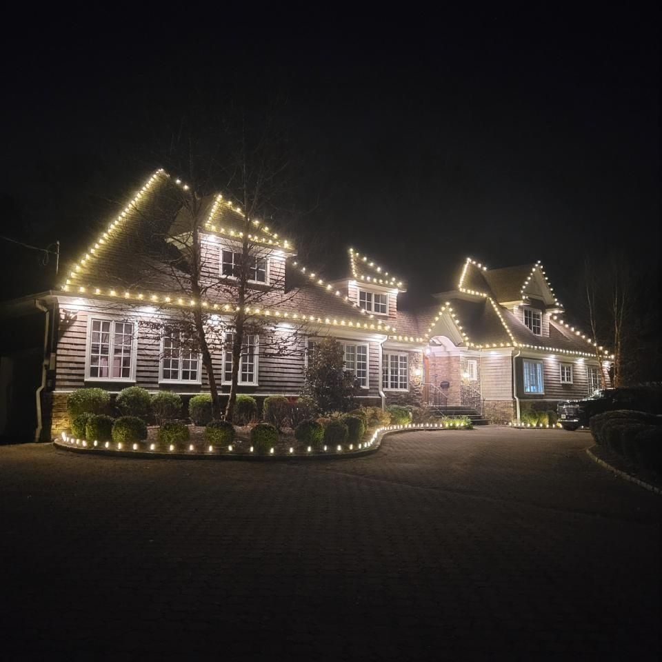 A large house at night outlined with warm white holiday lights and lit windows.