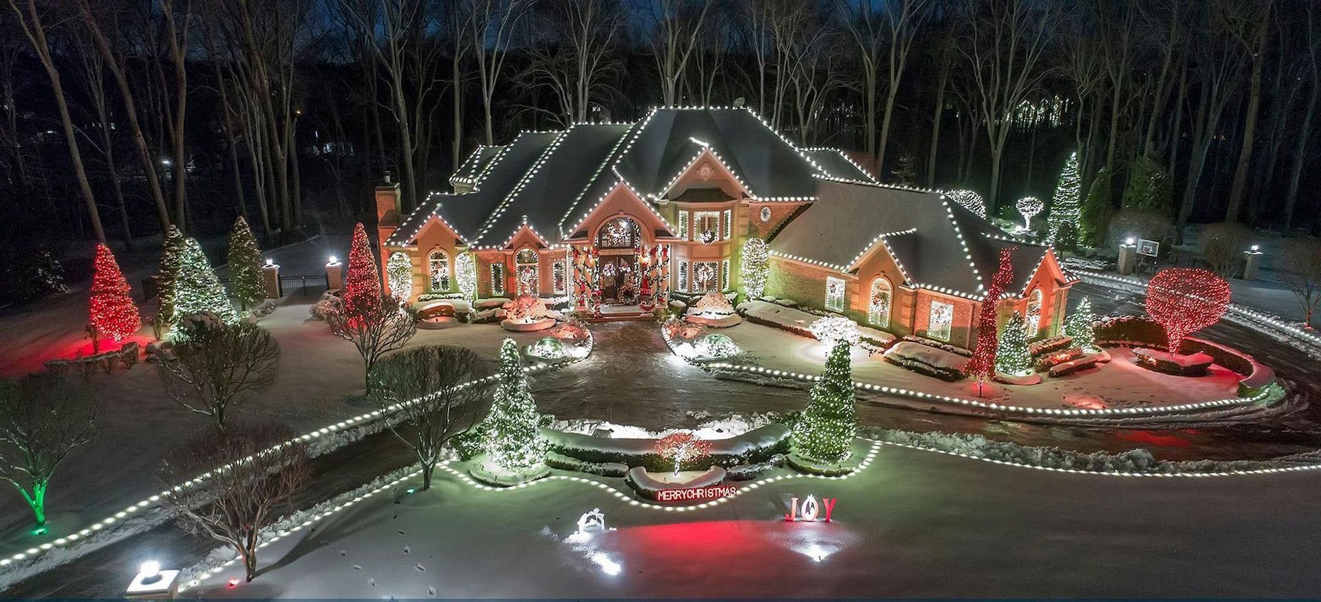 Large house decorated with red, green, and white Christmas lights at night, with a lit circular driveway and trees.