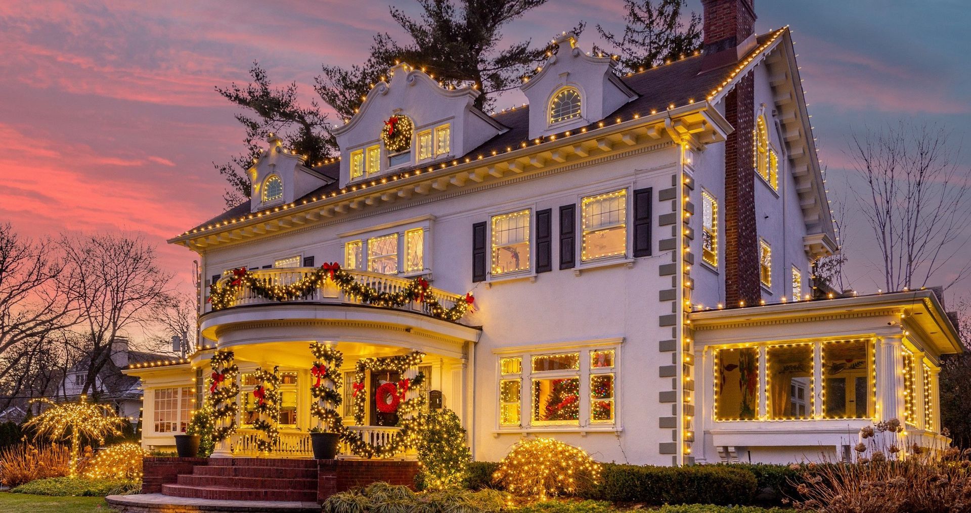 Festively lit Victorian house at dusk with glowing windows, garlands, and a pink sunset sky