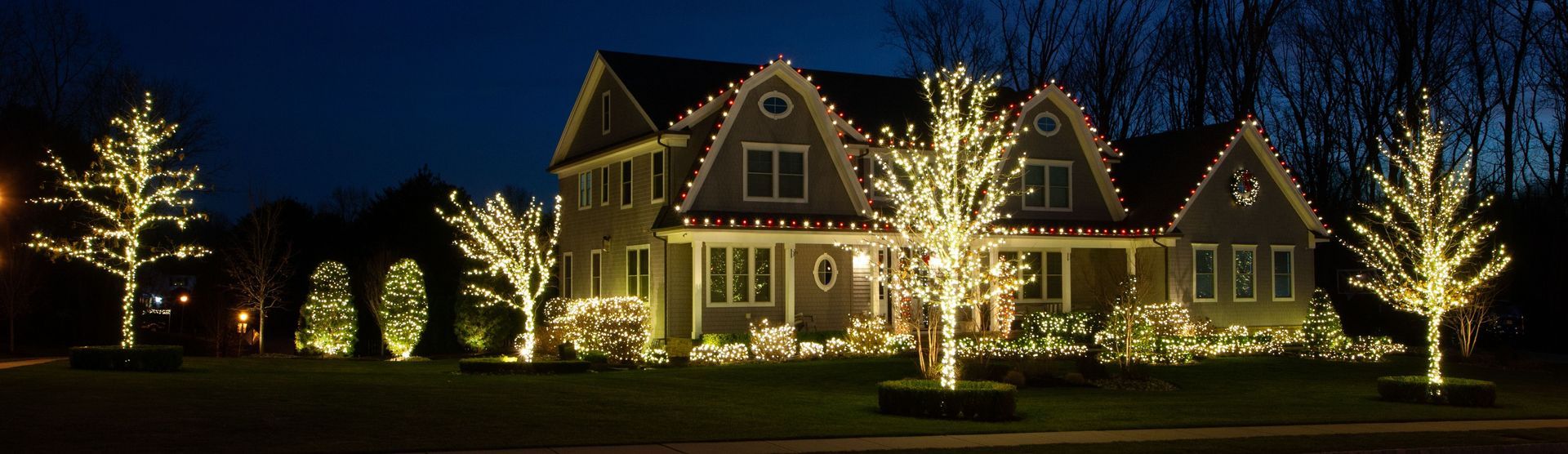 A warmly lit house decorated with glowing white Christmas lights at night, surrounded by illuminated trees.
