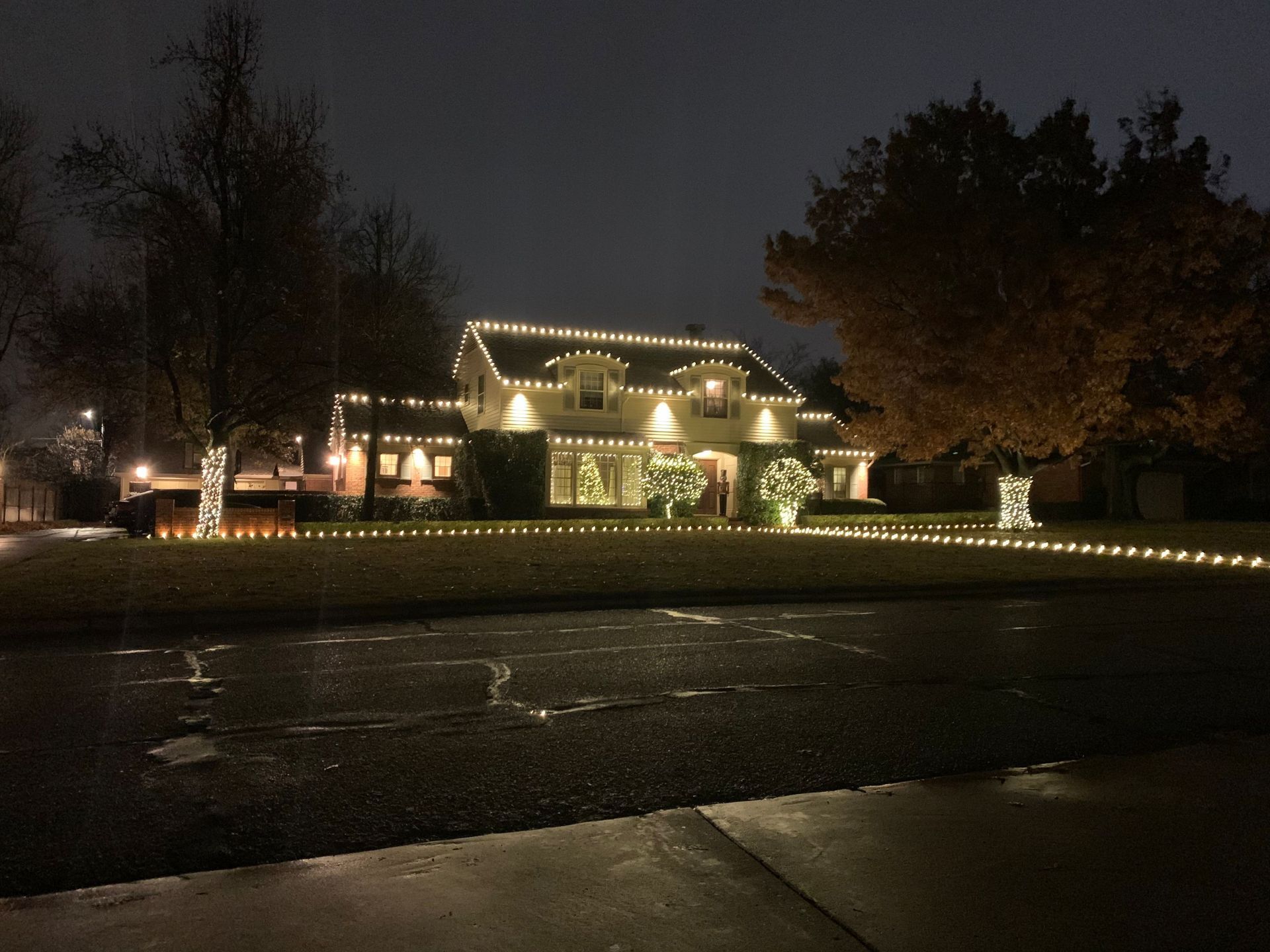 A house lit with warm white string lights at night, with a dark wet road in the foreground.