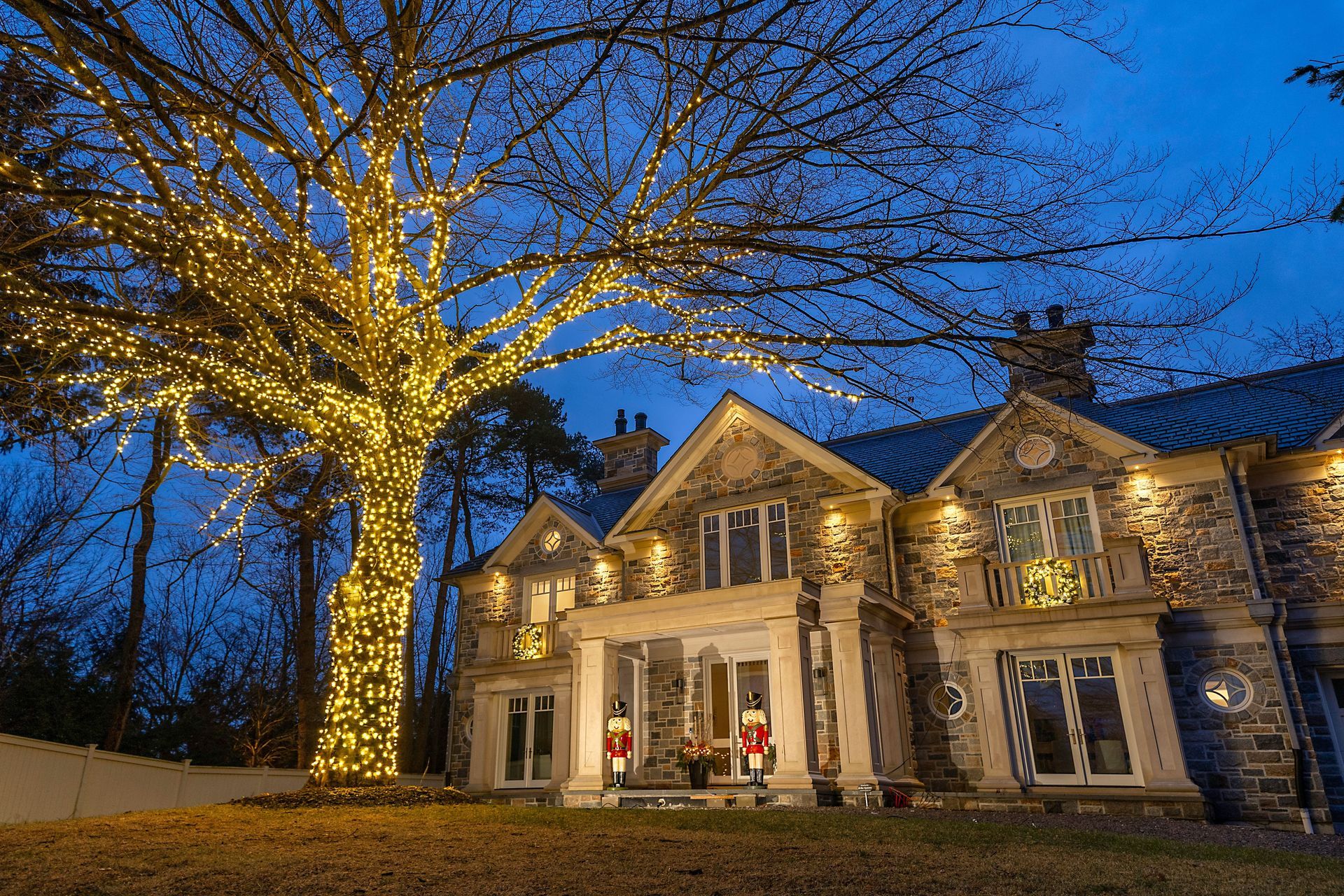 Large stone house lit warmly at dusk, with a tree wrapped in white lights in the front yard.
