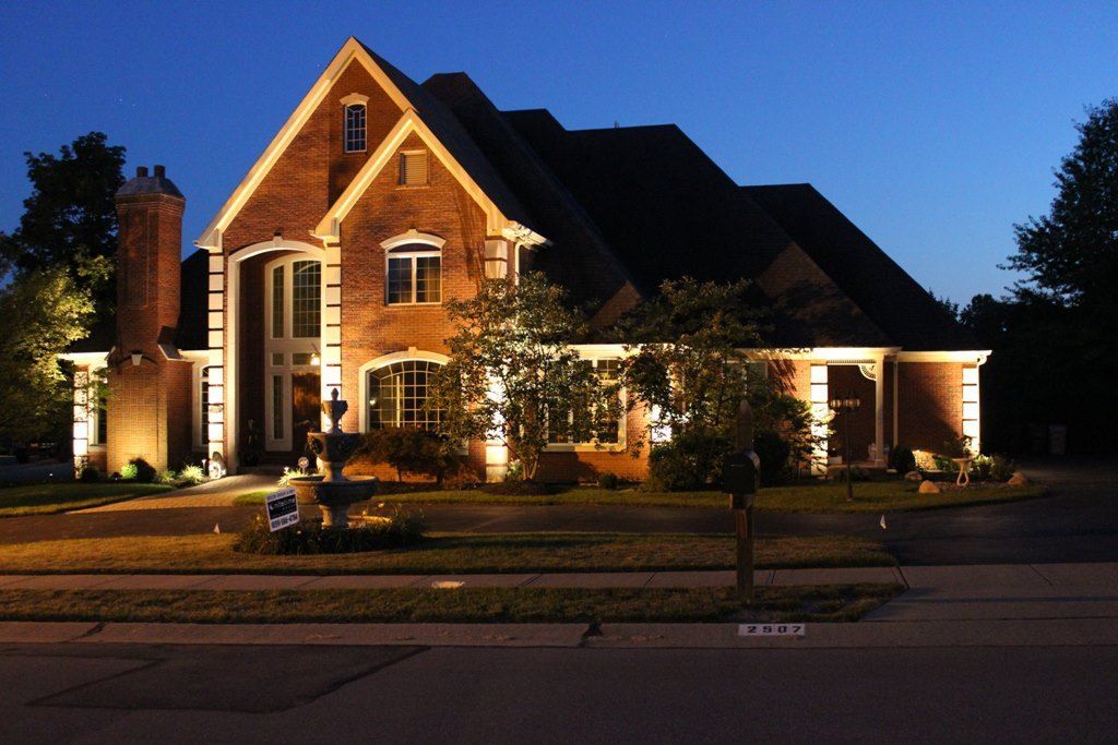 Brick house with warm porch lights at dusk, front lawn and driveway visible