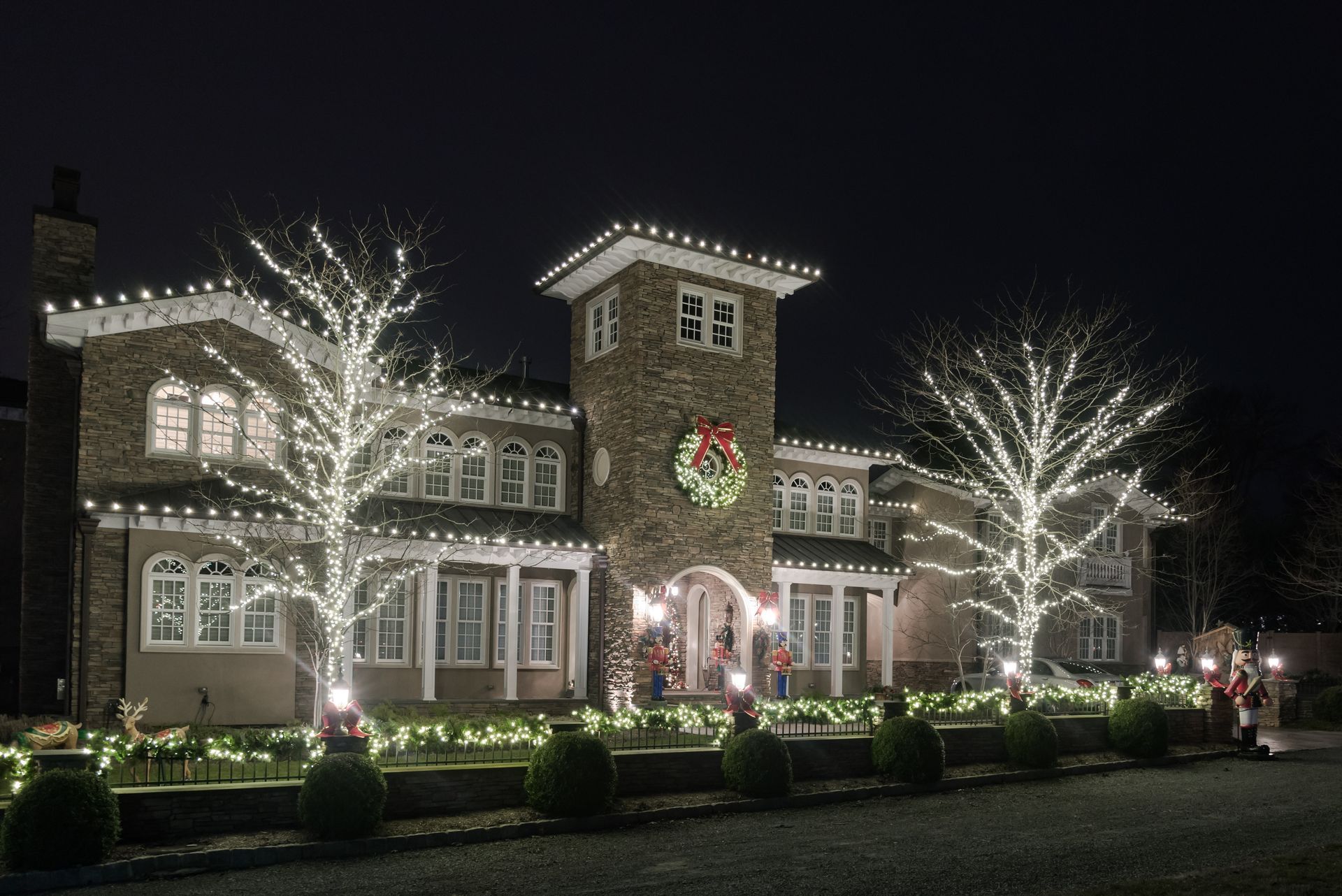 Large house decorated with white holiday lights, glowing wreaths, and lit trees at night