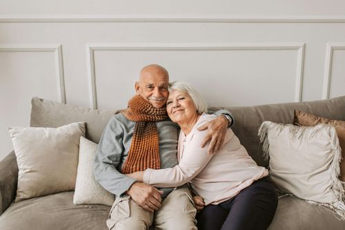 Couple cuddling on a gray couch in a bright living room, smiling and embracing.