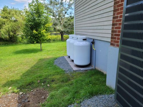 White propane tank beside a house, set on gravel in a grassy yard.
