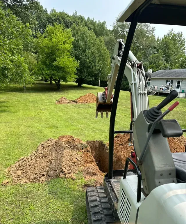 Mini excavator digging in a grassy yard, piles of dirt nearby. Trees and a house in the background.