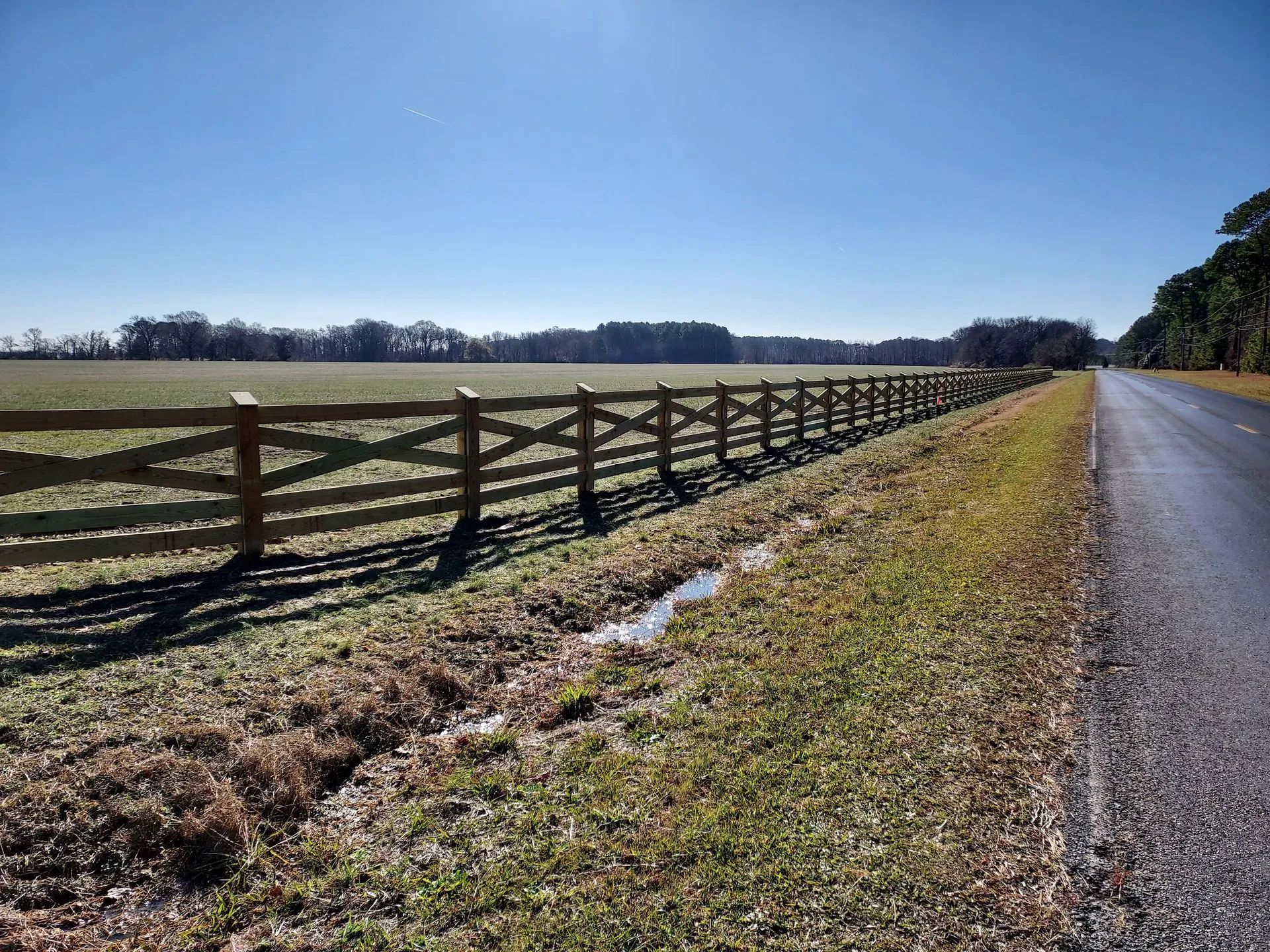 Wooden fence along a road, field beyond, bright sky, sun high.