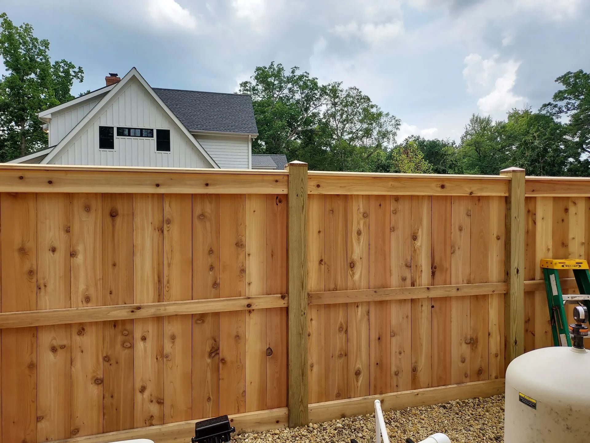 Wooden privacy fence in a backyard with a house and trees behind it under a partly cloudy sky