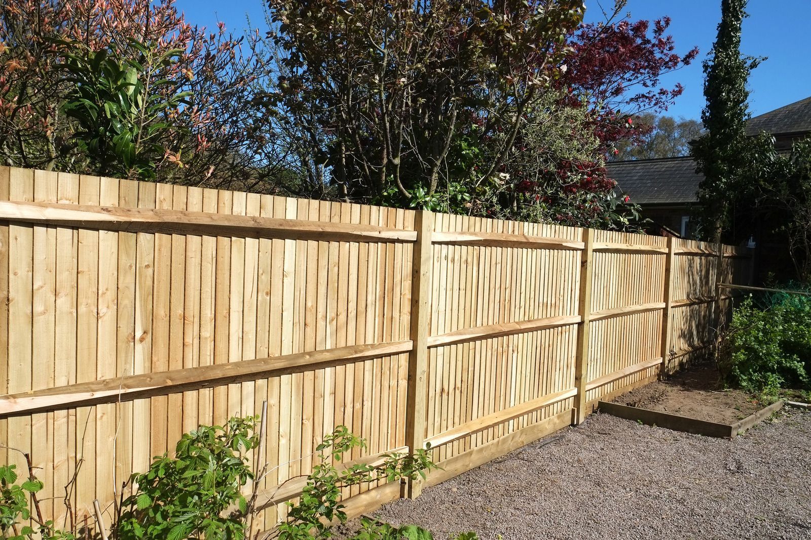 Wooden fence along a gravel path with trees and bushes behind it under a sunny blue sky