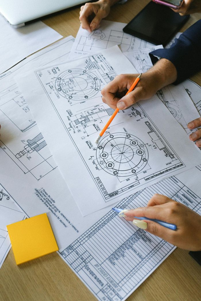 Hands reviewing technical drawings with pencils and a yellow notepad on a desk