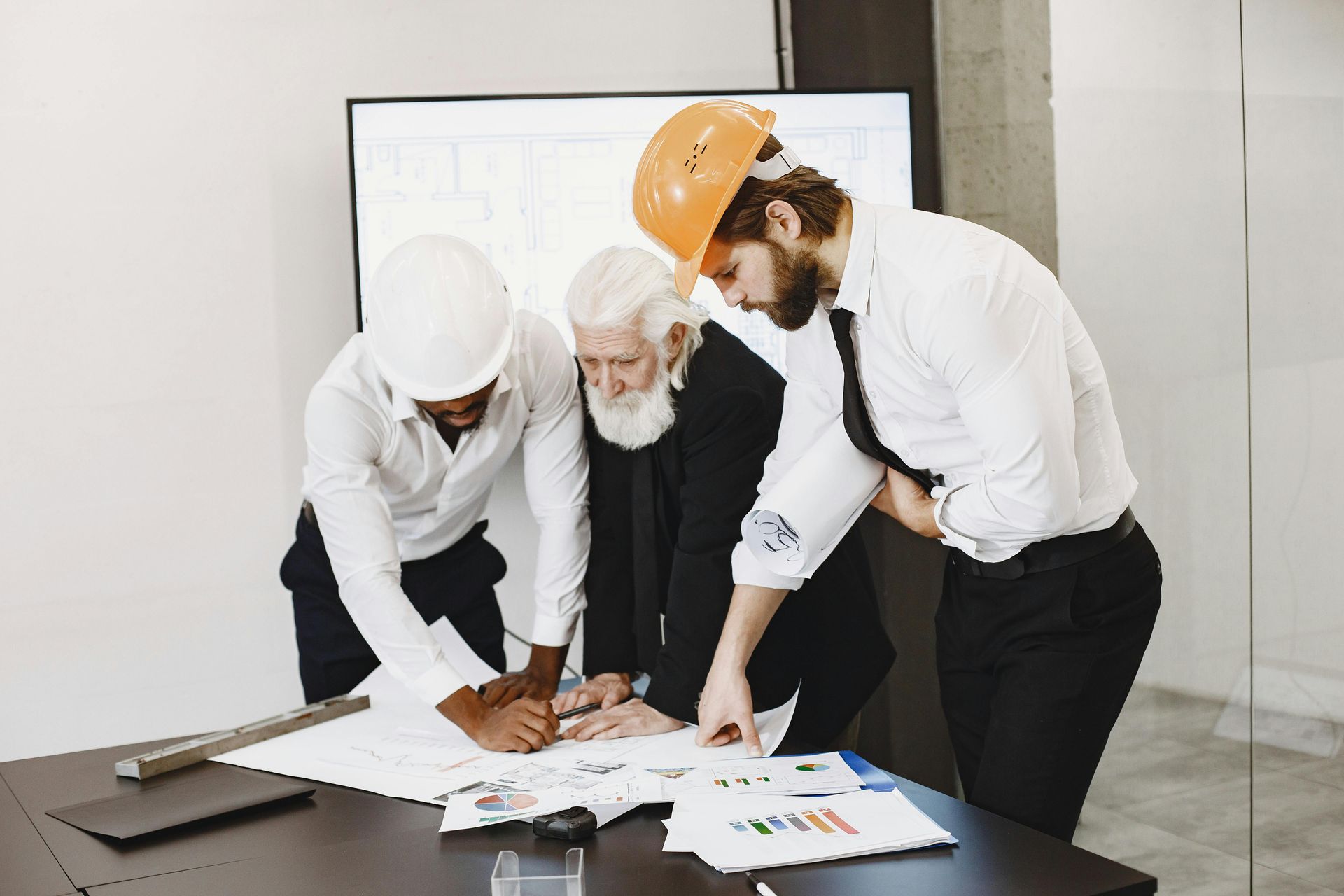 Three workers in hard hats review plans at a table in an office, pointing at documents.