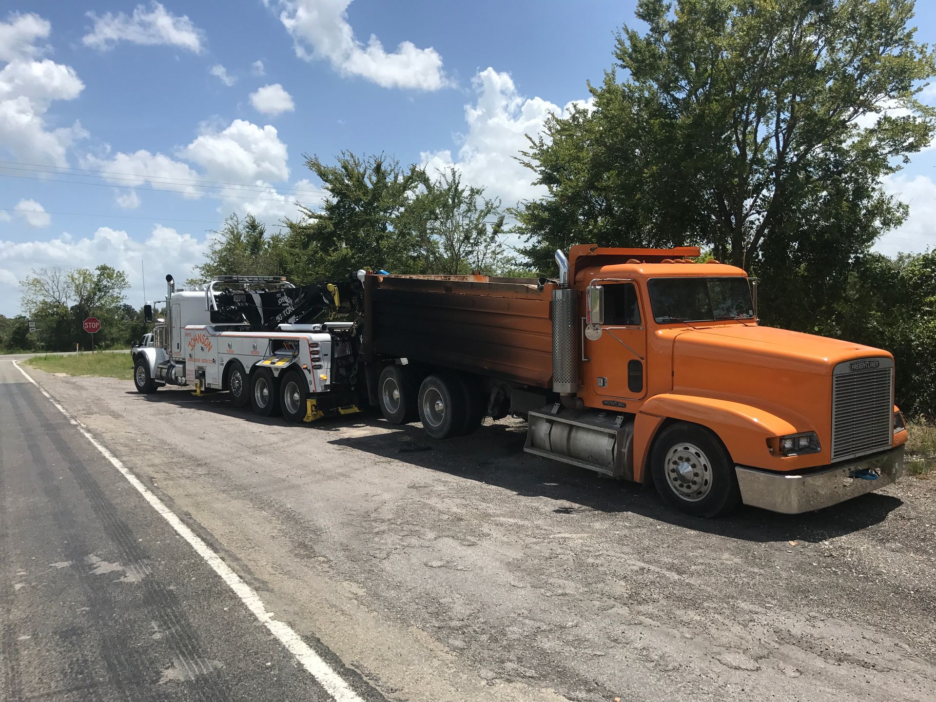 A white tow truck towing an orange dump truck on the side of a road on a sunny day.