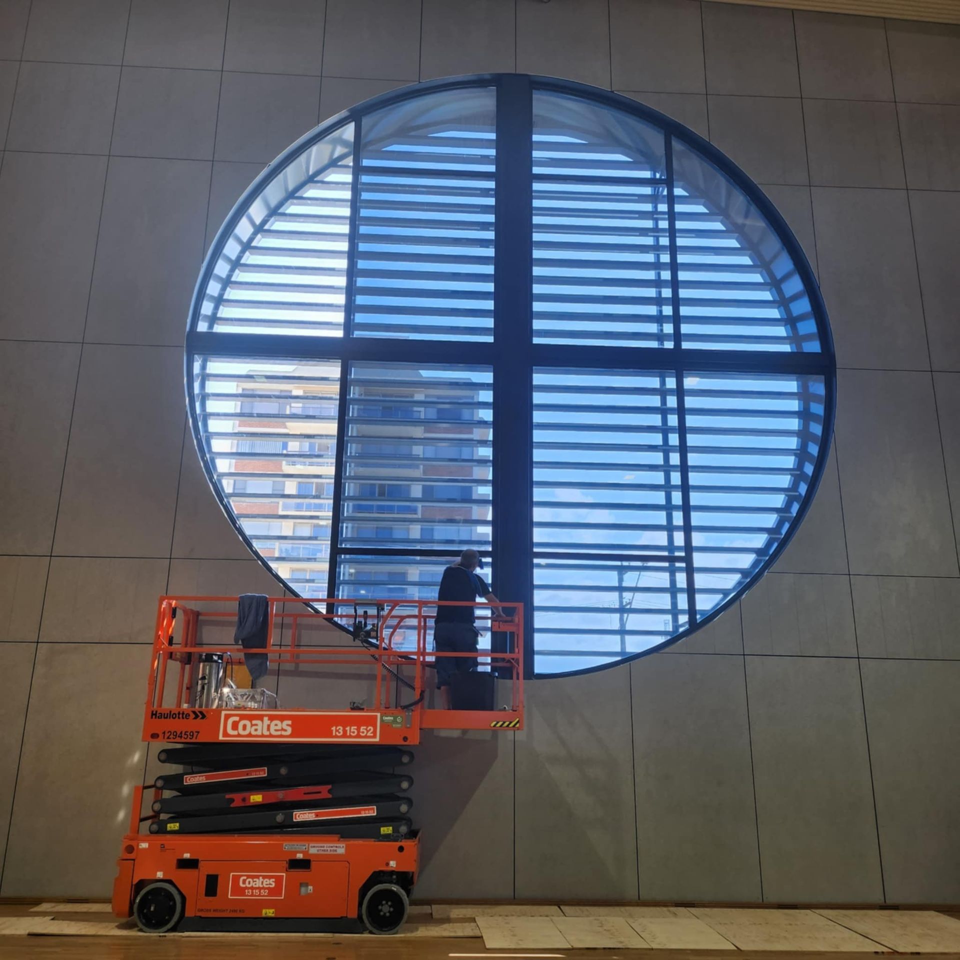 Person on a lift working on a large, round window with horizontal slats. Orange lift, building exterior.