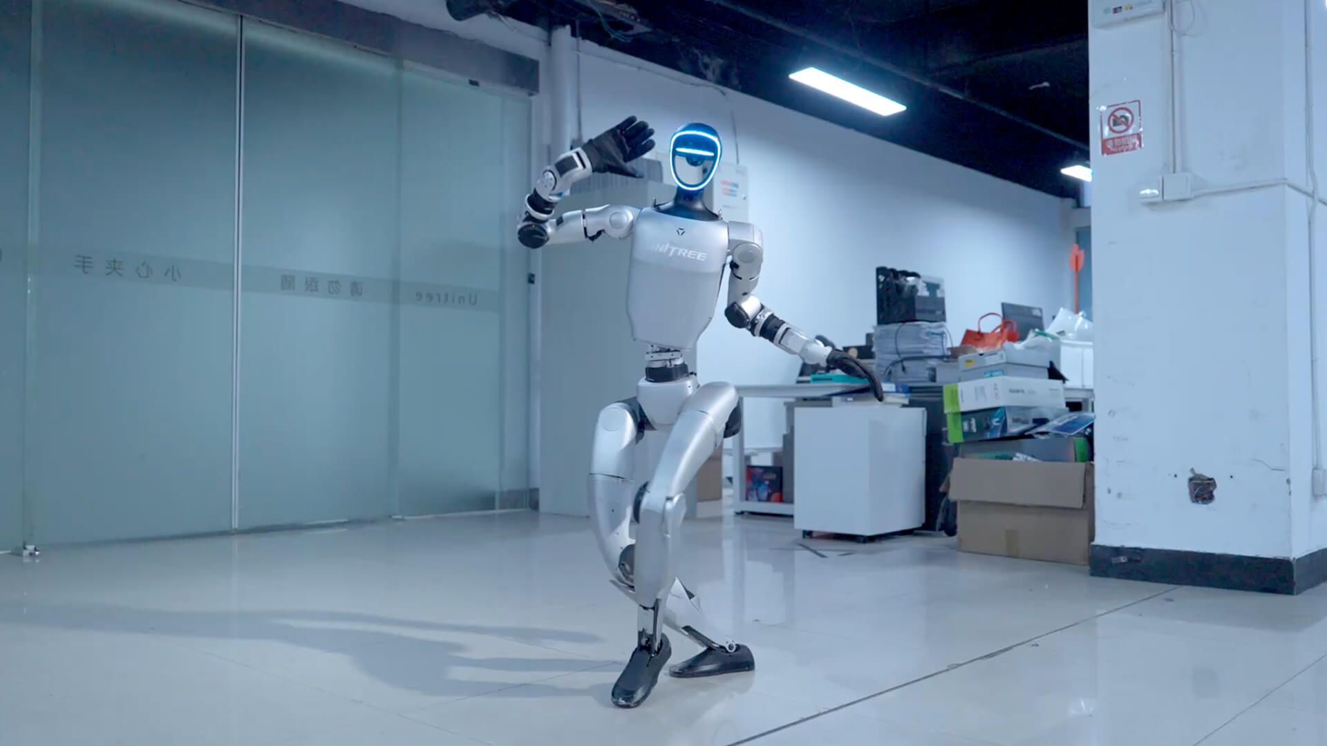 Robot, silver and blue, dances in a white-walled room with a desk.