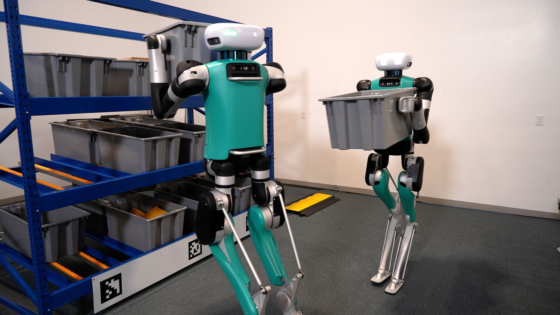 Two legged robots in a warehouse setting, carrying gray bins. One robot near a rack, another walking.