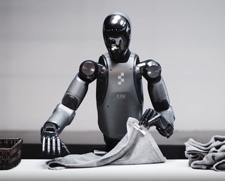 Robot folding a gray towel on a white table. Another basket and folded towels are nearby.