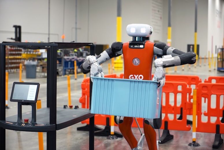 Robot in orange and gray carrying a blue crate in a warehouse setting with barriers.