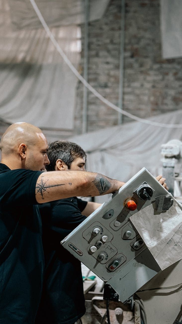 Two people operating machinery in a workshop; one points to a control panel.