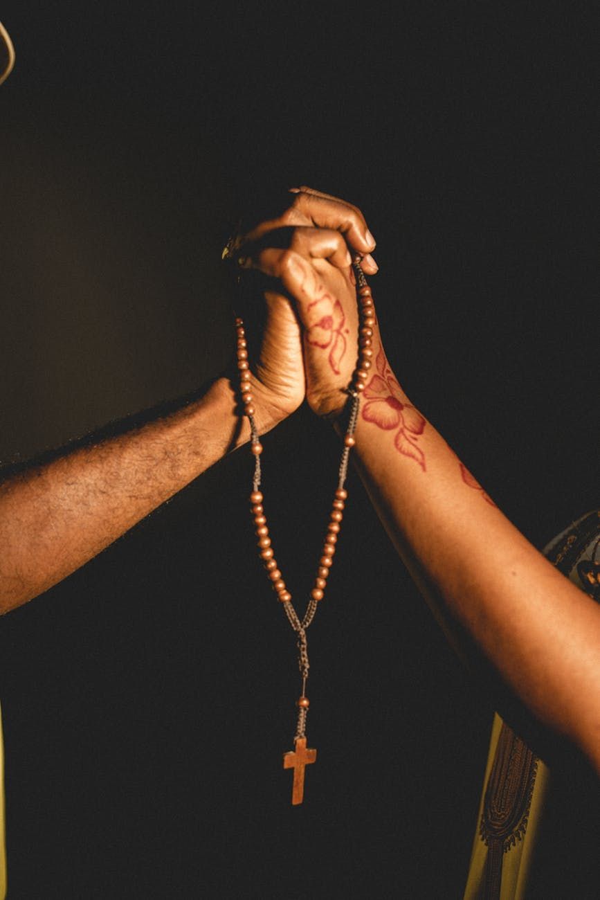 Hands clasped, holding a rosary. One wrist has a henna design. Dark background, warm lighting.