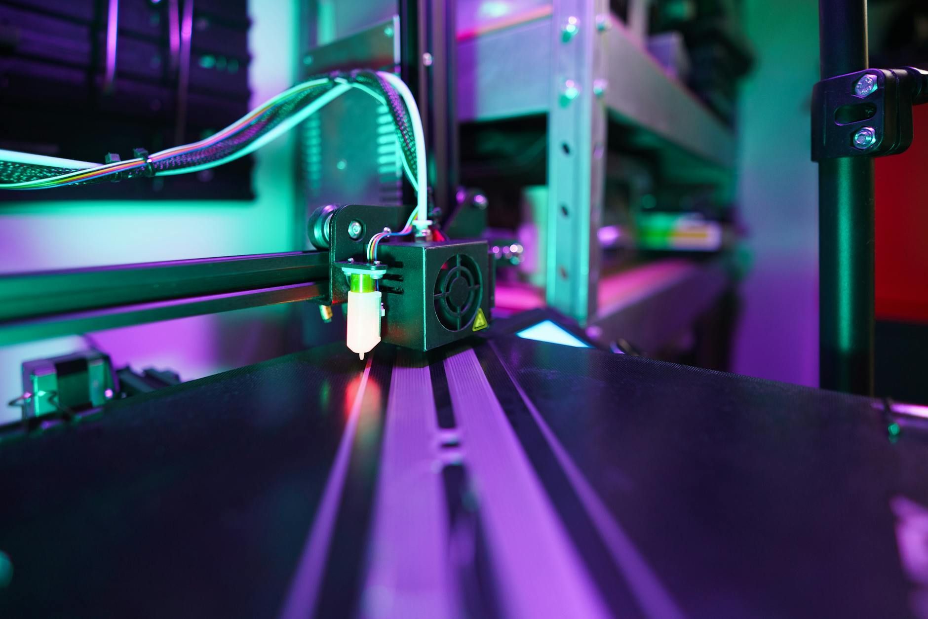 A 3D printer head depositing melted filament onto a black print bed. Purple and green lighting.