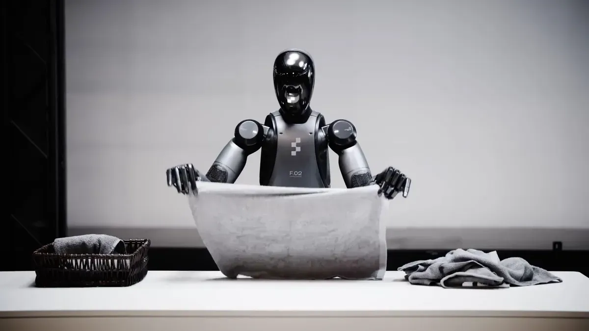 Robot folds a towel at a table, with a basket and laundry. Gray and black tones.