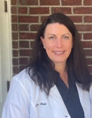 A professional in a white lab coat standing in front of a brick wall, smiling.