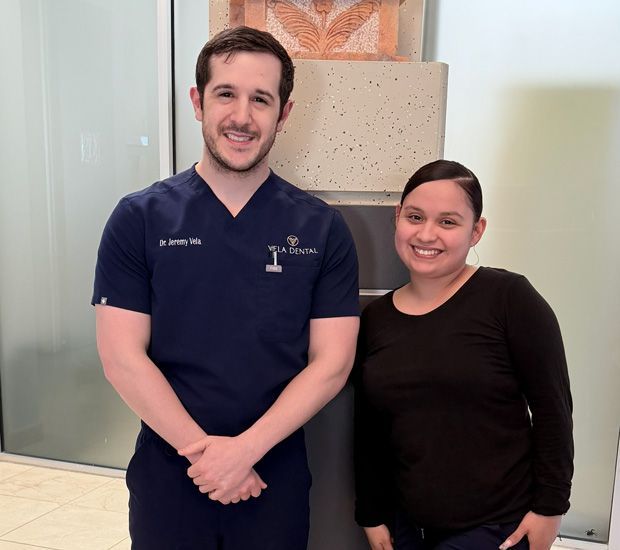 Two smiling people standing in a bright office lobby, one in navy scrubs and one in a black top.