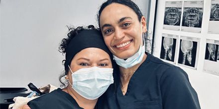 Two smiling masked people standing together in a room with photos on the wall